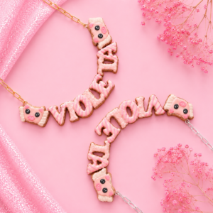 Collares personalizados de cerdito con alas kawaii con nombres decorados en rosa, uno con cadena dorada y otro con cadena plateada.